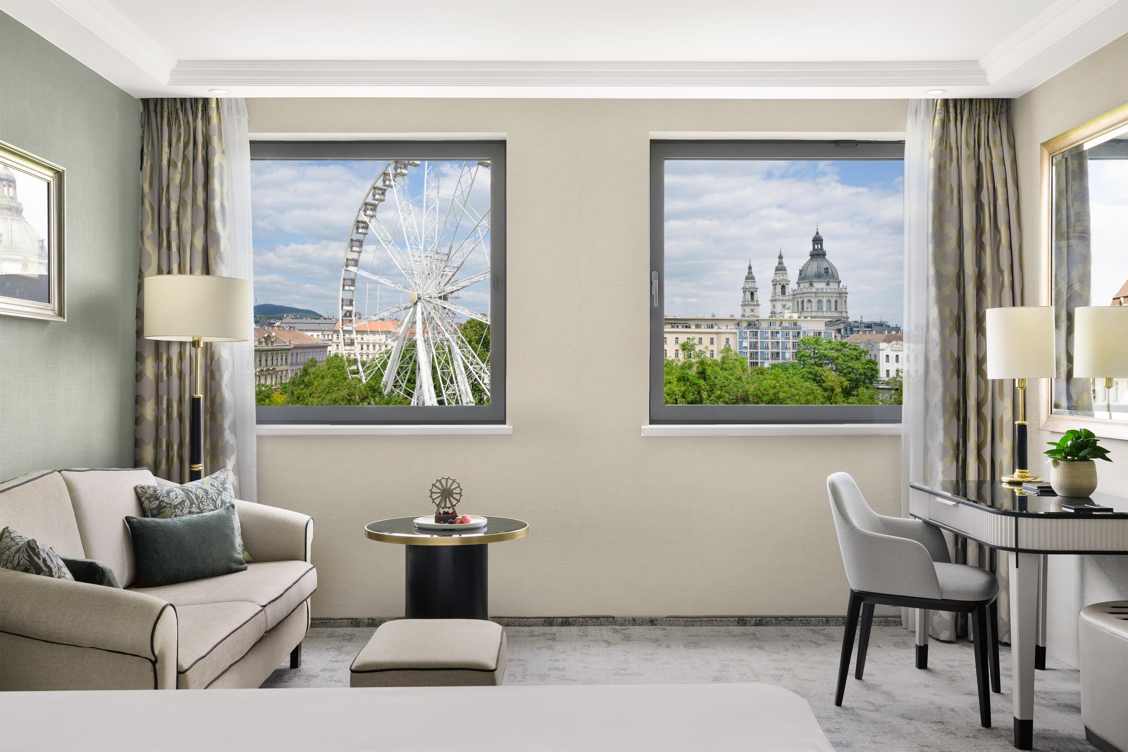 Habitación de hotel con cama, sofá, escritorio, dos ventanas con vistas a la ciudad y una mesita con un objeto decorativo.