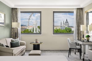A hotel room with a bed, a sofa, a desk, two windows with city views, and a small table with a decorative item. - Kempinski Hotel Corvinus Budapest (Budapest)
