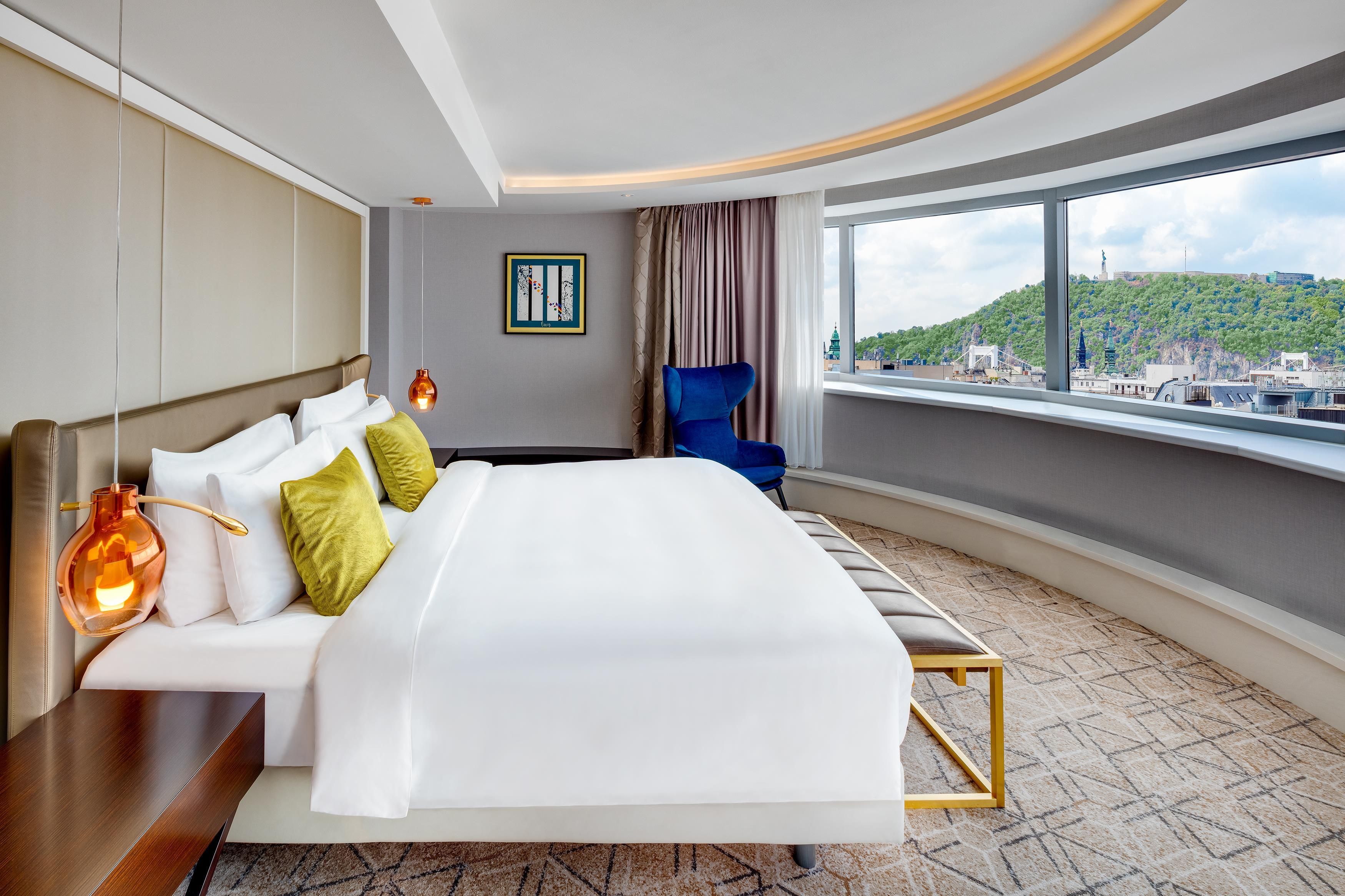 Una habitación de hotel con una cama grande, una silla, vistas a la ciudad y una obra de arte enmarcada en la pared.