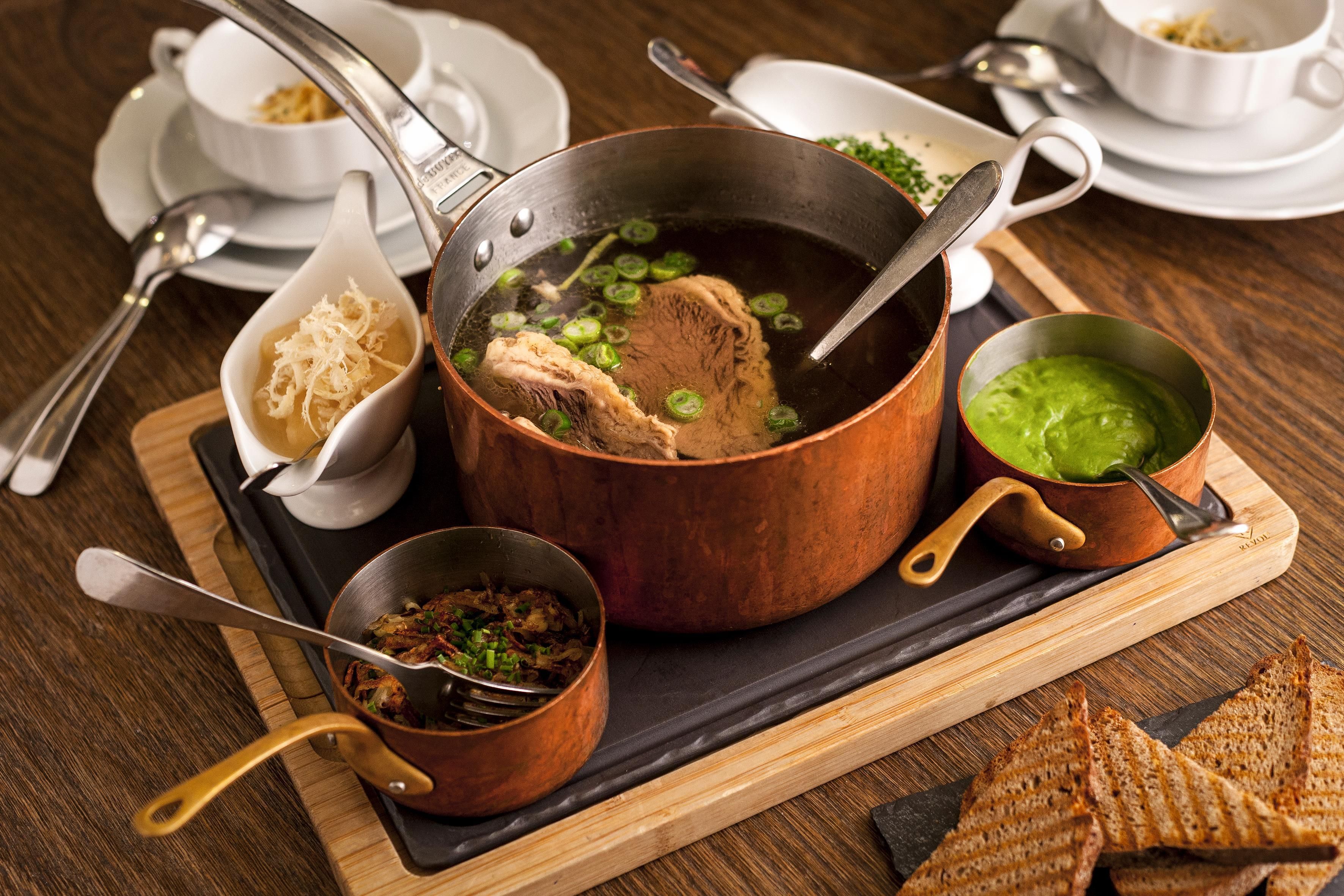 Een eettafel met een grote pan soep, een kom groene saus en een kleine schaal gesneden ingrediënten.