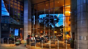 A modern hotel with a glass facade, outdoor seating area, and a visible bar named "Blue Fox The Bar". - Kempinski Hotel Corvinus Budapest (Budapest)