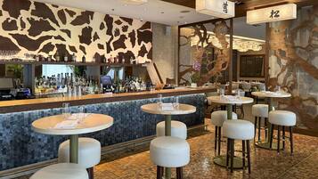 Uma área de bar com balcão, banquetas e um espaço de estar com mesas redondas.