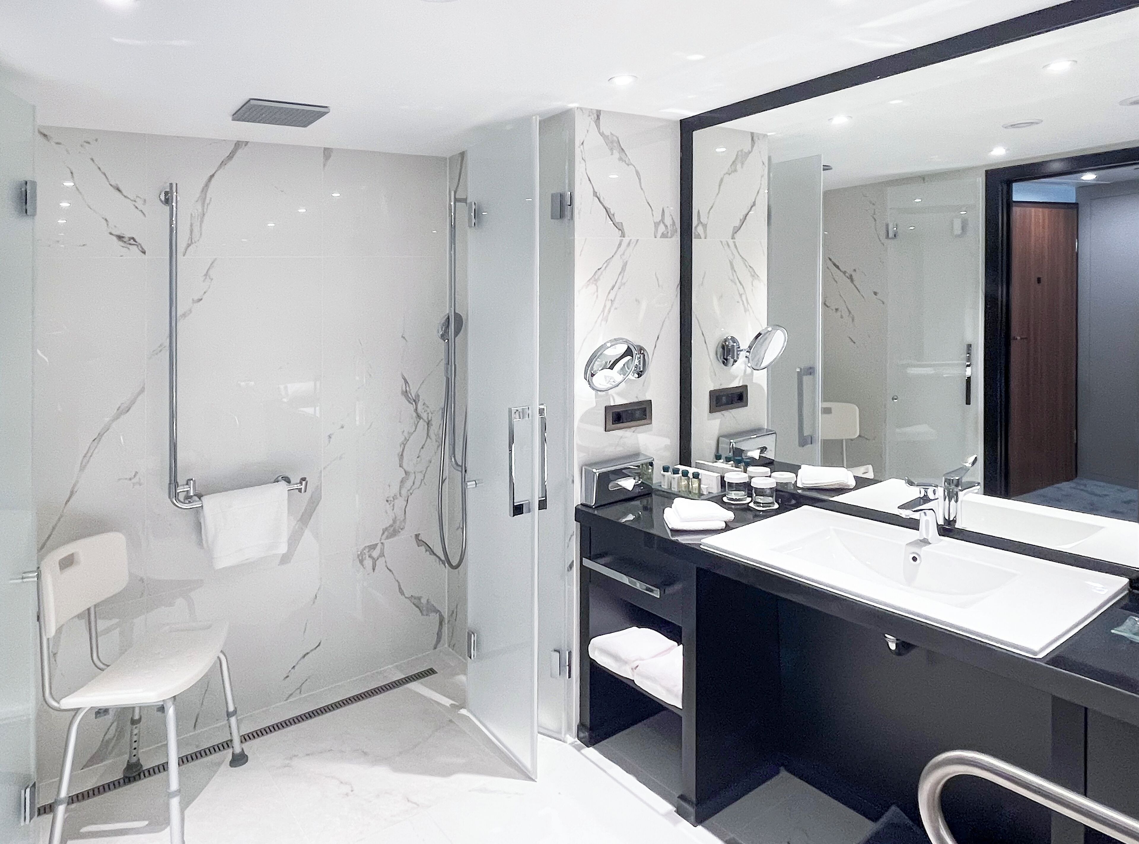 Un baño moderno con ducha envuelta en cristal, pared de mármol blanco, espejo grande y tocador negro con dos lavamanos.