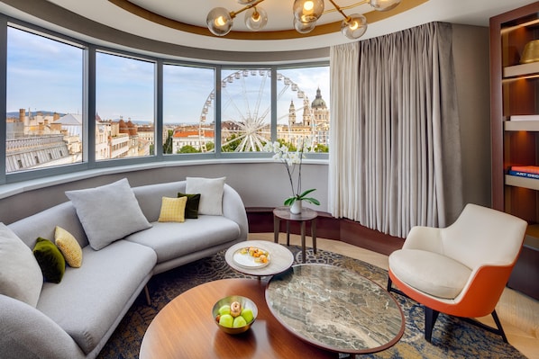 Una sala moderna con sofá, sillón y una mesita de centro, vista a un paisaje urbano y una noria, además de una estantería con libros.