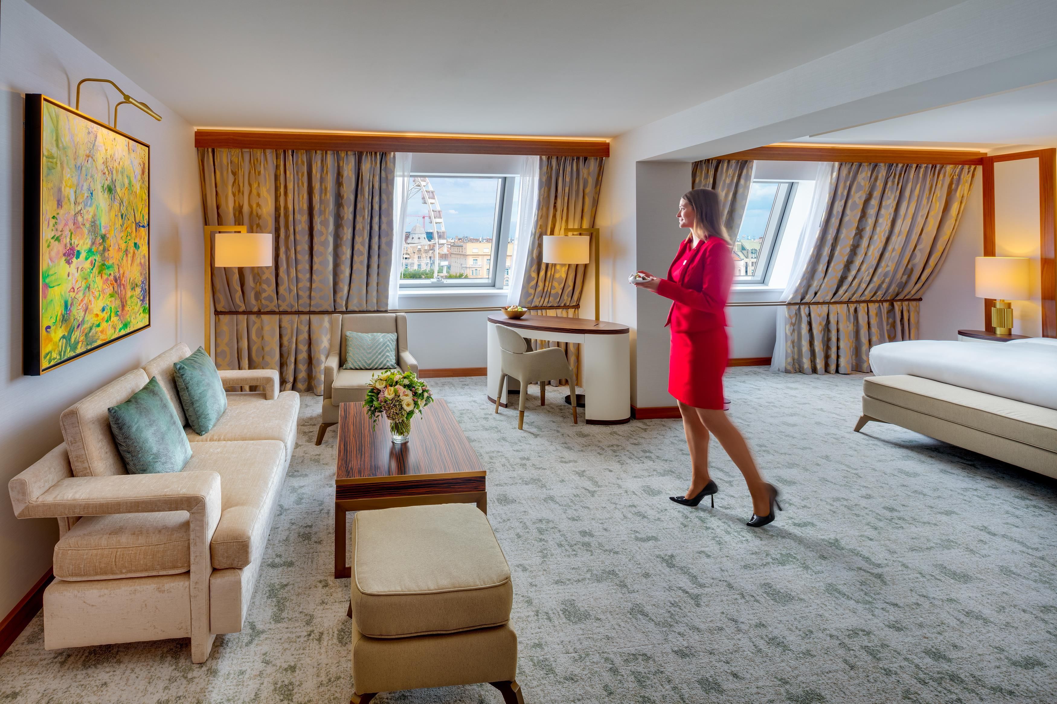 Habitación de hotel con sofá, sillón, escritorio y vistas a la ciudad a través de amplias ventanas.