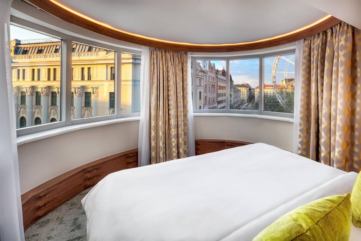 Hotellihuone, jossa on suuri sänky, kaksi ikkunaa verhoilla sekä näkymä kaupungin rakennuksiin ja näköalarattaaseen.
