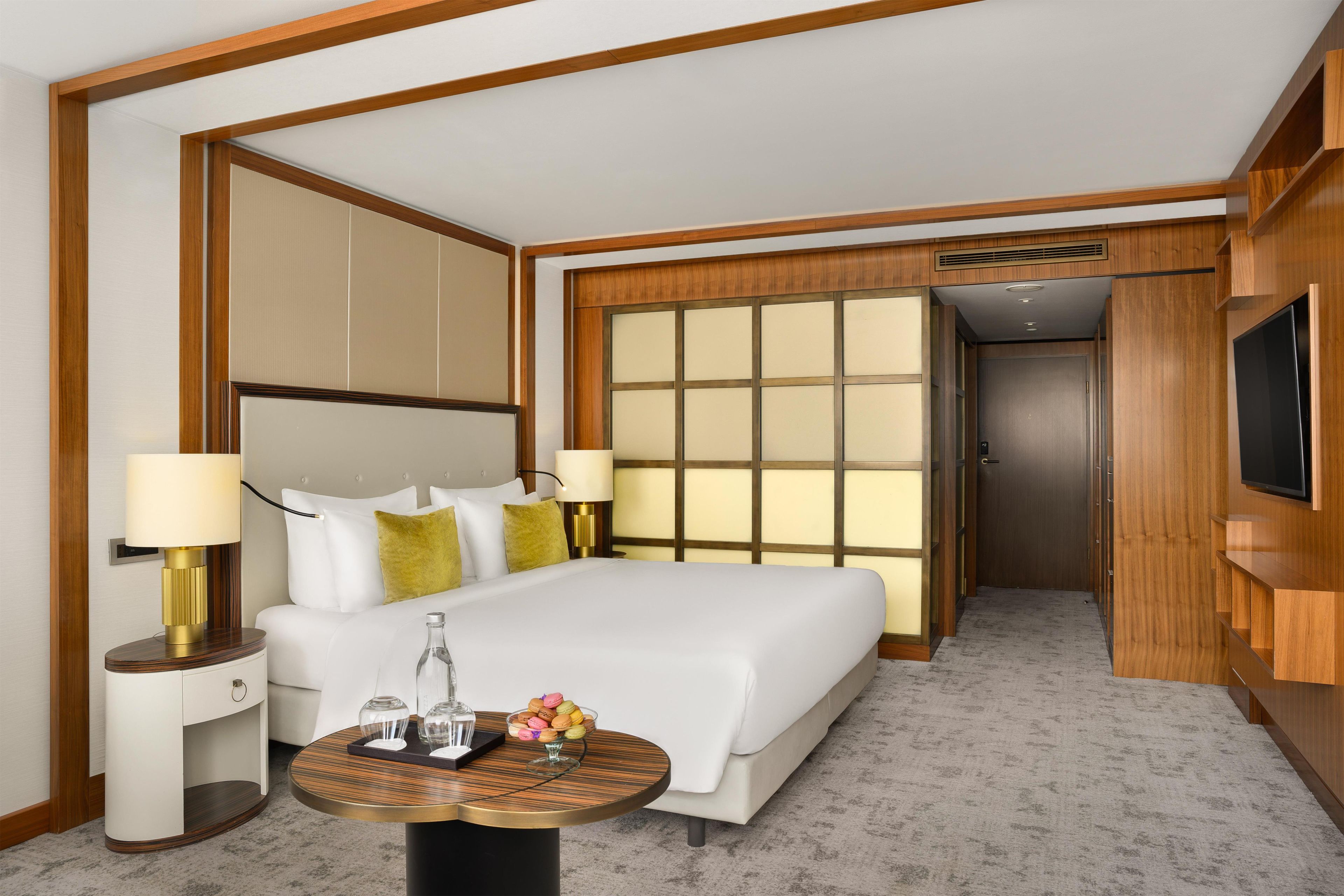 A hotel room with a bed, bedside tables, a television, and a view of a corridor.