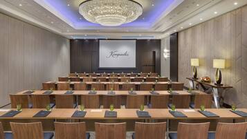 A conference room with rows of wooden tables and chairs, a large screen displaying "Kempinski", and a chandelier.