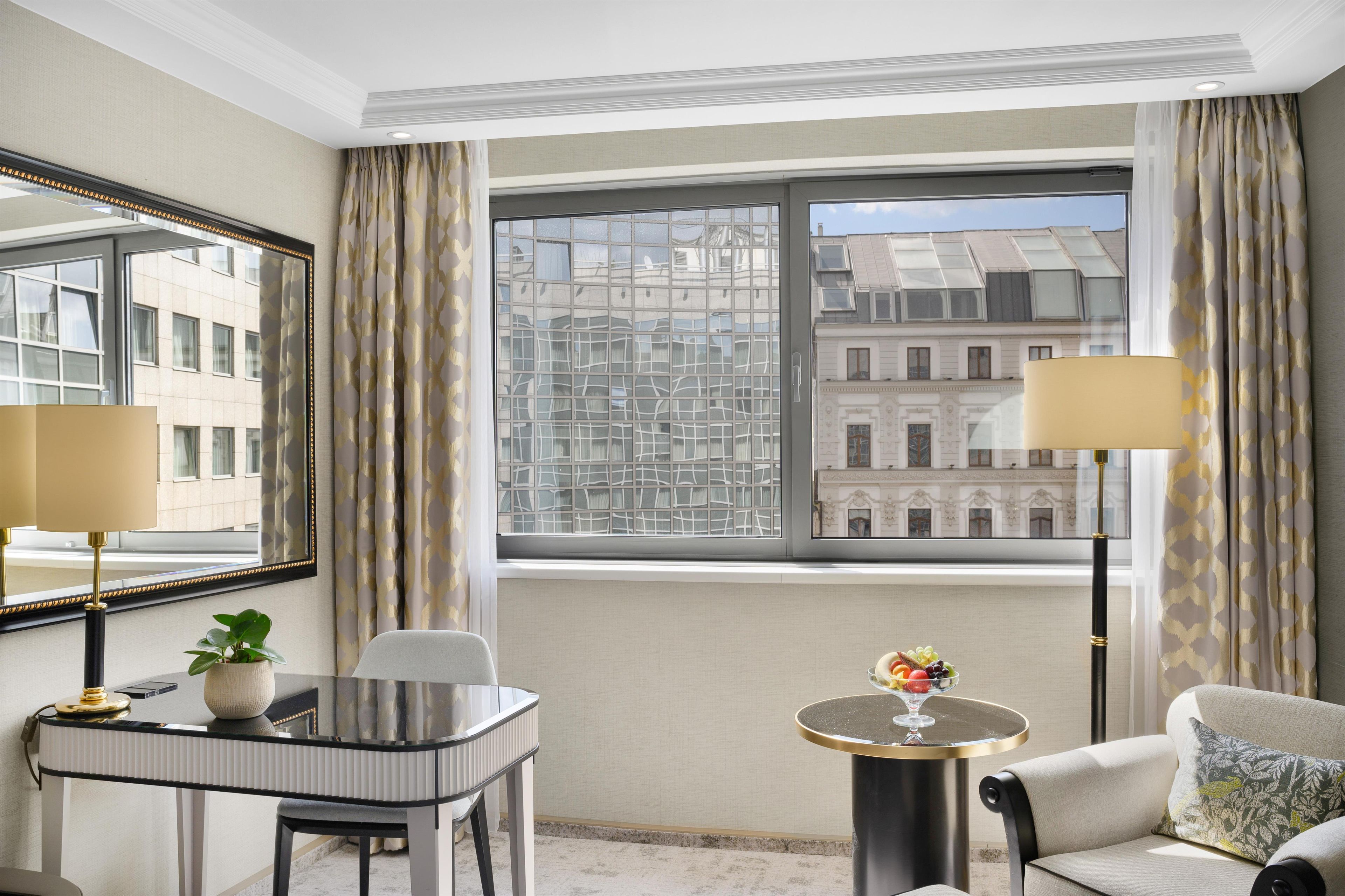 A hotel room with a large window, a desk with a chair, a small round table with a fruit bowl, a sofa, and a mirror.