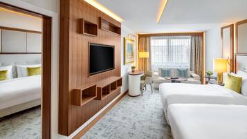 A hotel room with a large flat-screen TV, two beds, a sofa, a small table, and a window with curtains.
