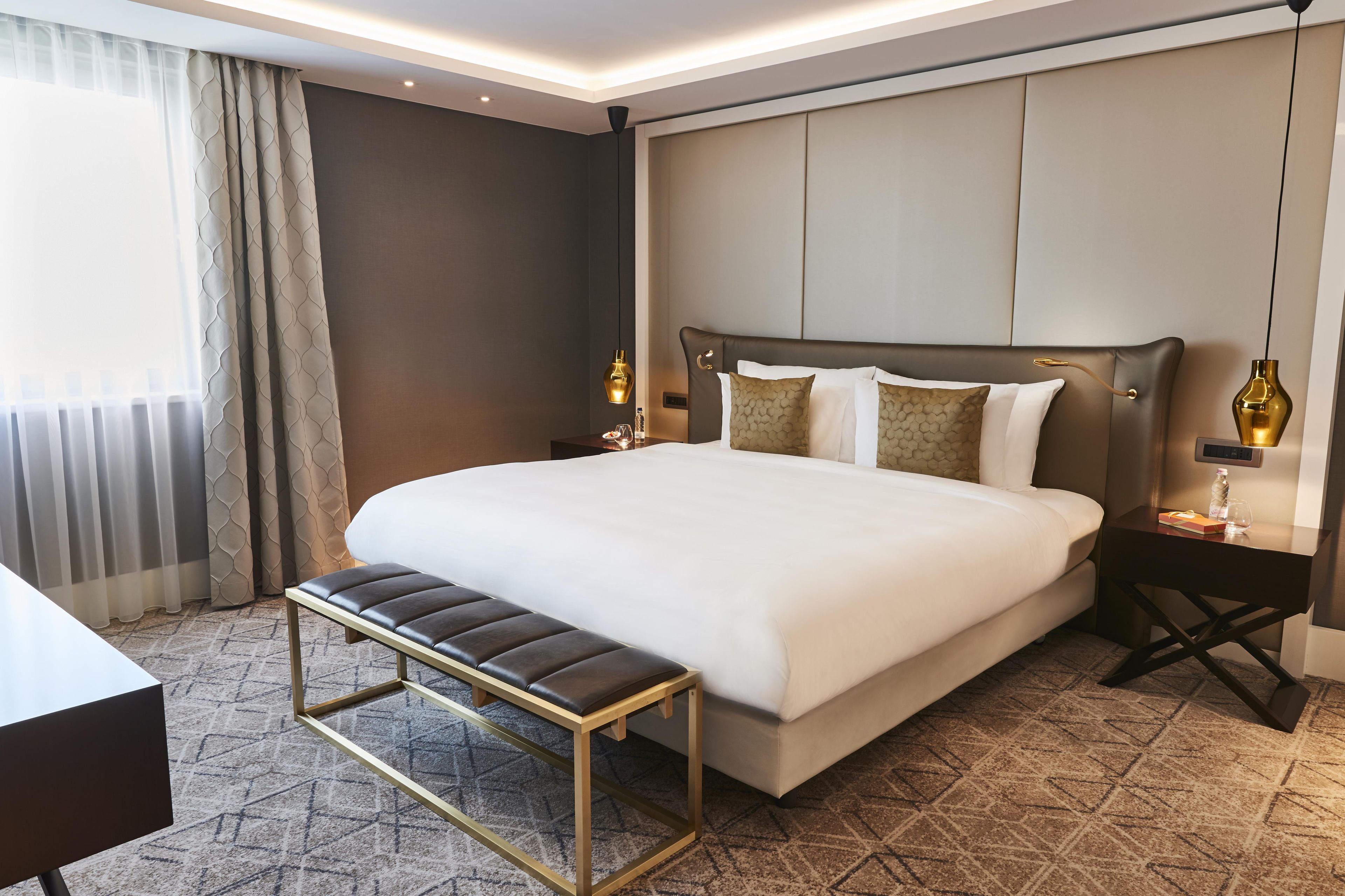 A hotel room with a large bed, a bench, a desk, and a nightstand with a lamp.