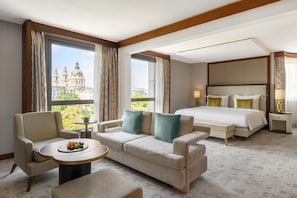 A hotel room with a large bed, a sofa, a small table, and a view of a historic building. - Kempinski Hotel Corvinus Budapest (Budapest)