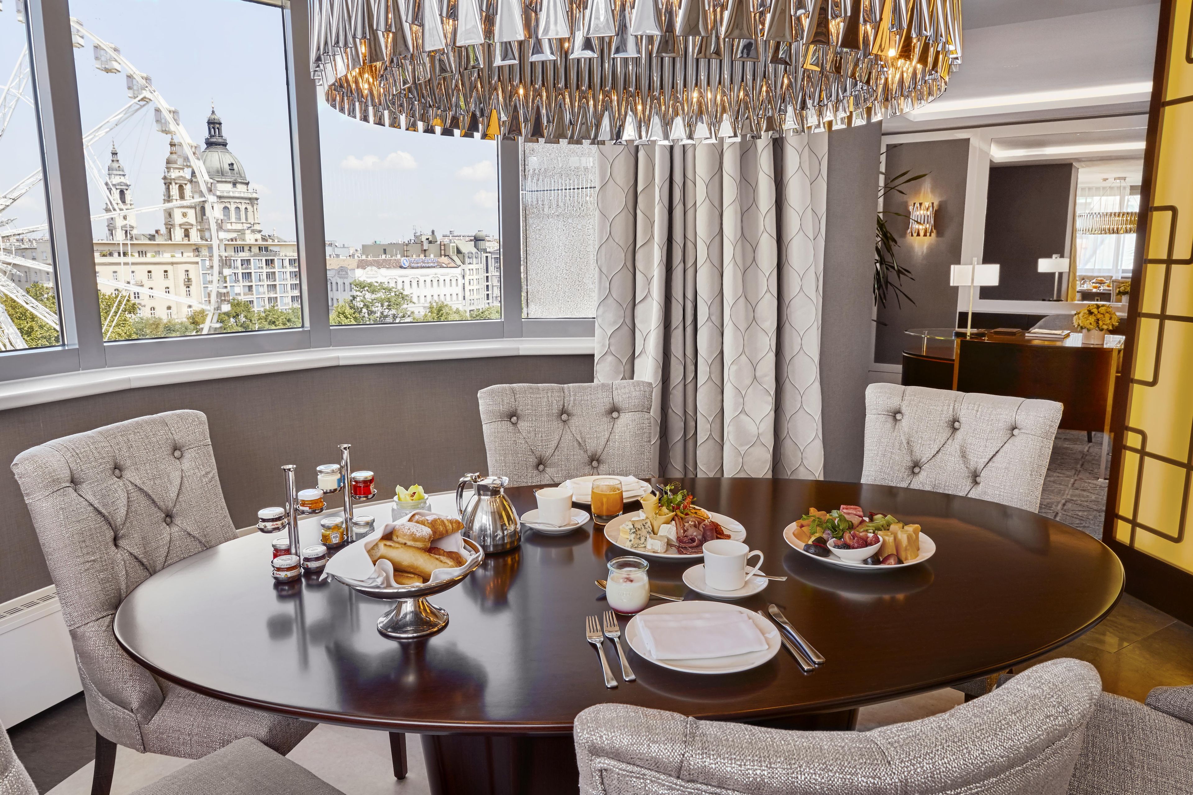 Comedor con una mesa redonda dispuesta para una comida, ventanas amplias con vista a un paisaje urbano y una lámpara de araña moderna.