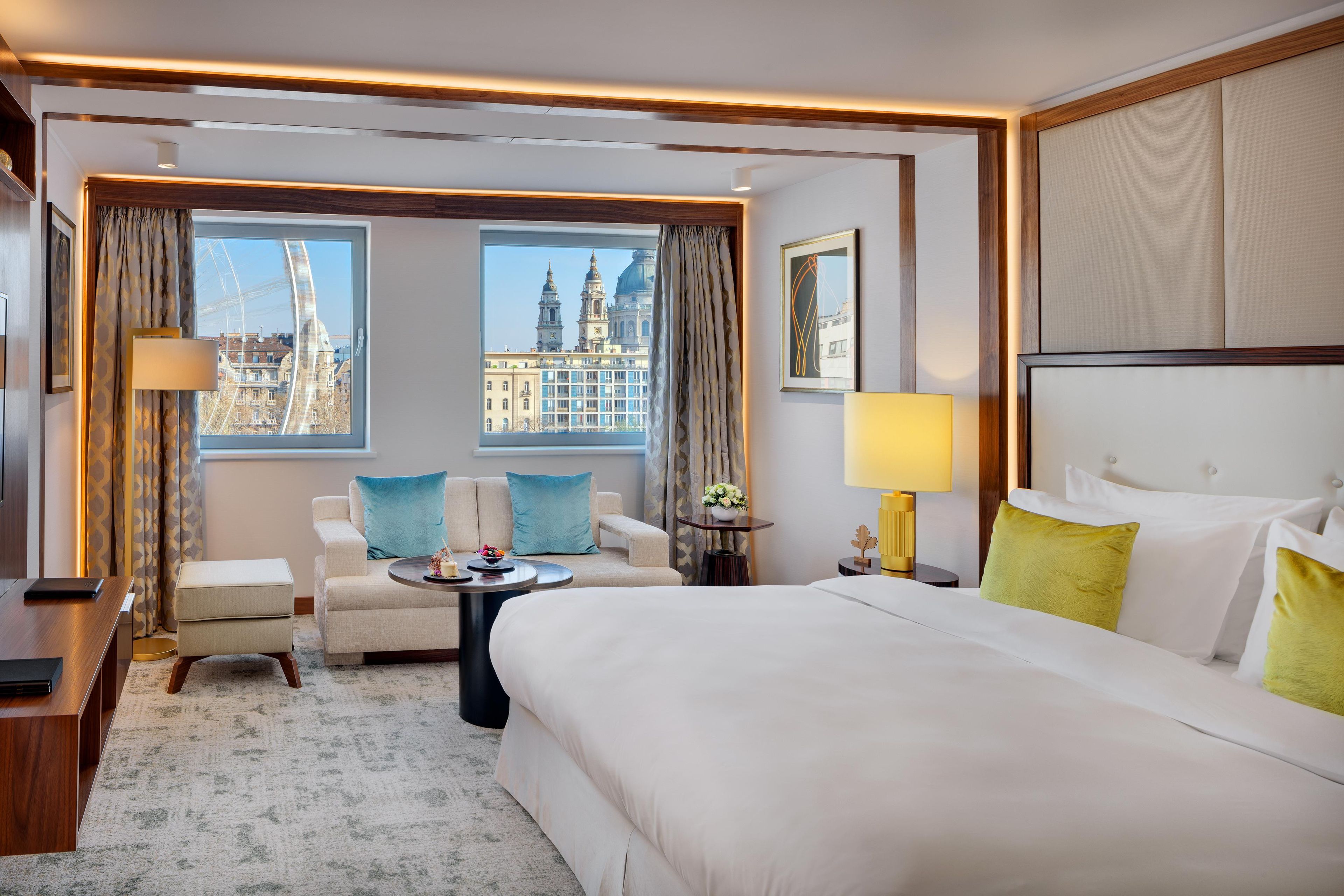 A hotel room with a large bed, two armchairs, a small table, and a view of city buildings through the windows.