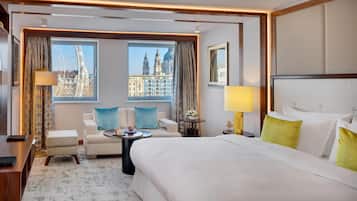 A hotel room with a large bed, two armchairs, a small table, and a view of city buildings through the windows.