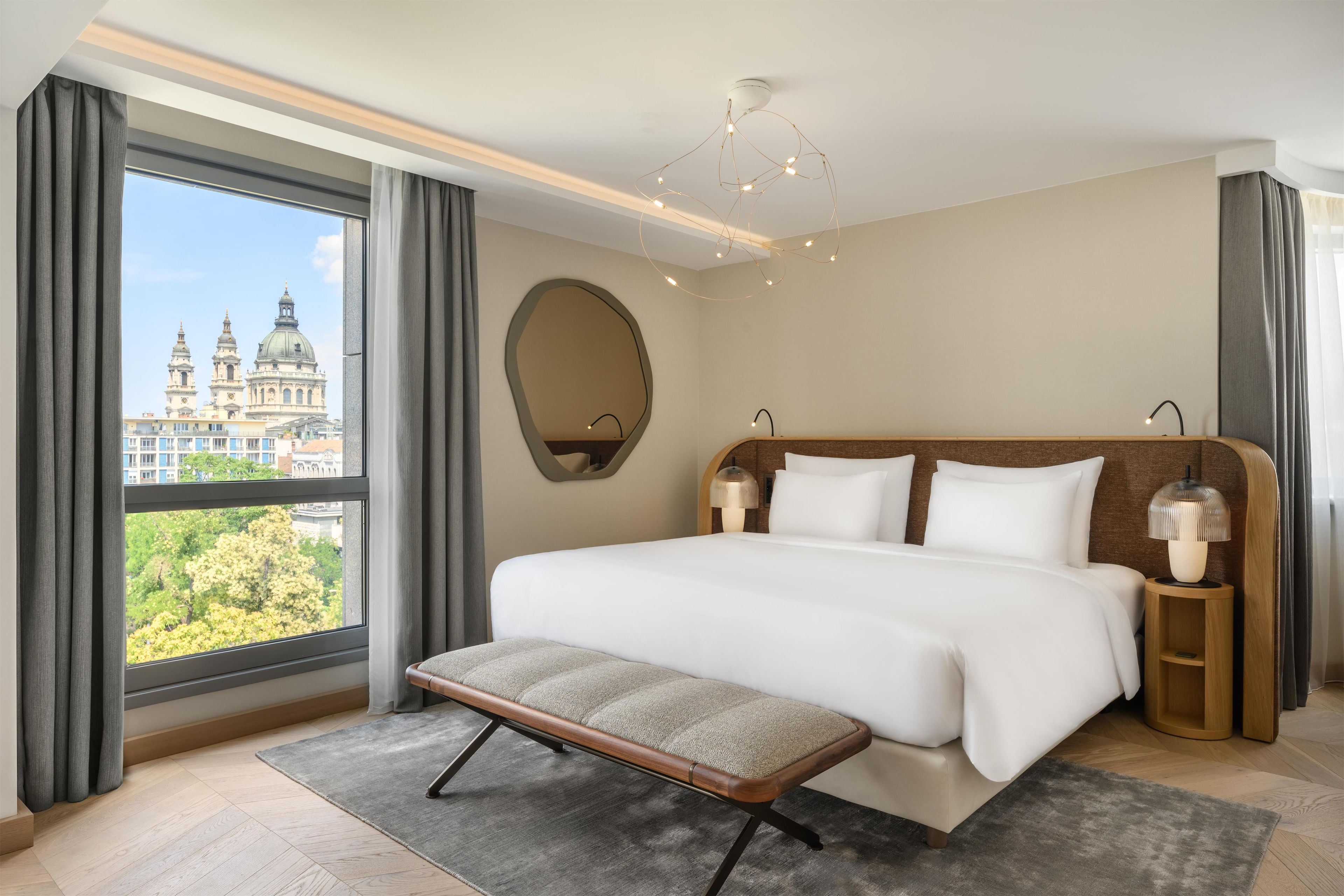 A hotel room with a large bed, a bench, a bedside table with a lamp, a mirror, and a view of a cityscape.