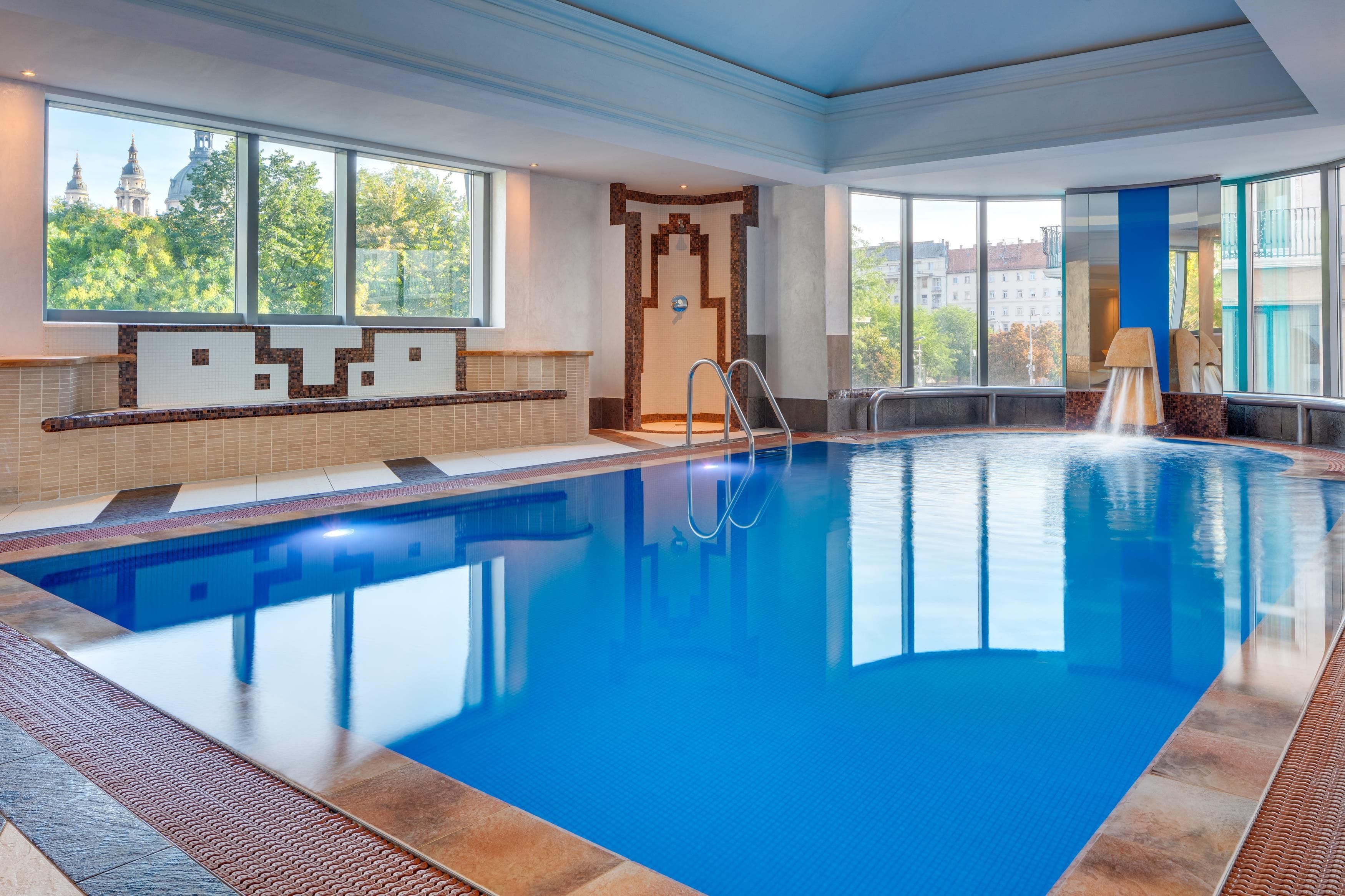 A large indoor swimming pool with clear blue water, a diving board, and a view of a cityscape through large windows.