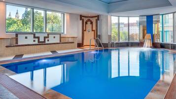 A large indoor swimming pool with clear blue water, a diving board, and a view of a cityscape through large windows.