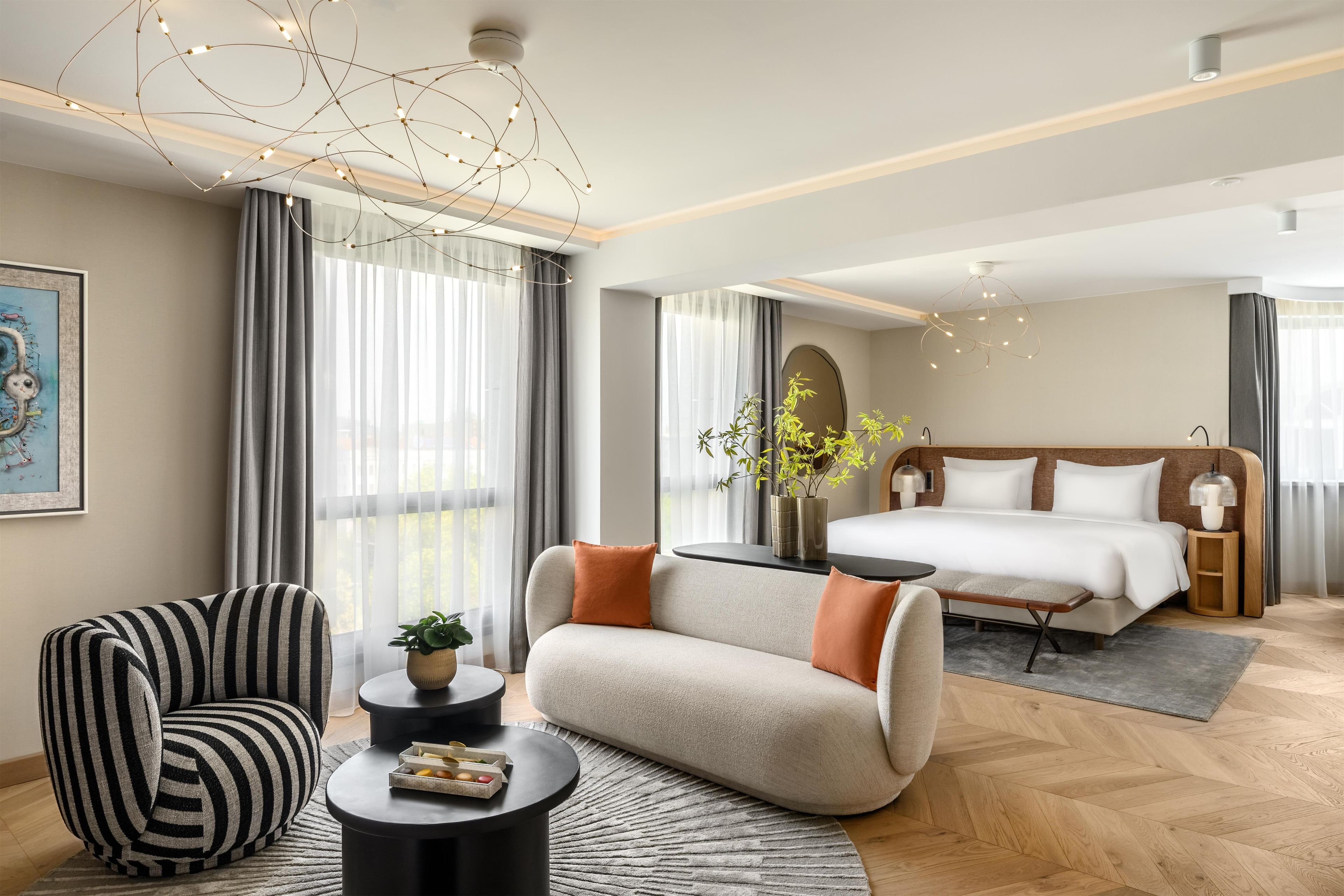 A modern hotel room with a sofa, armchair, round coffee table, and a bed with a wooden headboard.