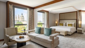 A hotel room with a large bed, a sofa, a small table with a tray of food, and a view of a historic building. - Kempinski Hotel Corvinus Budapest (Budapest)