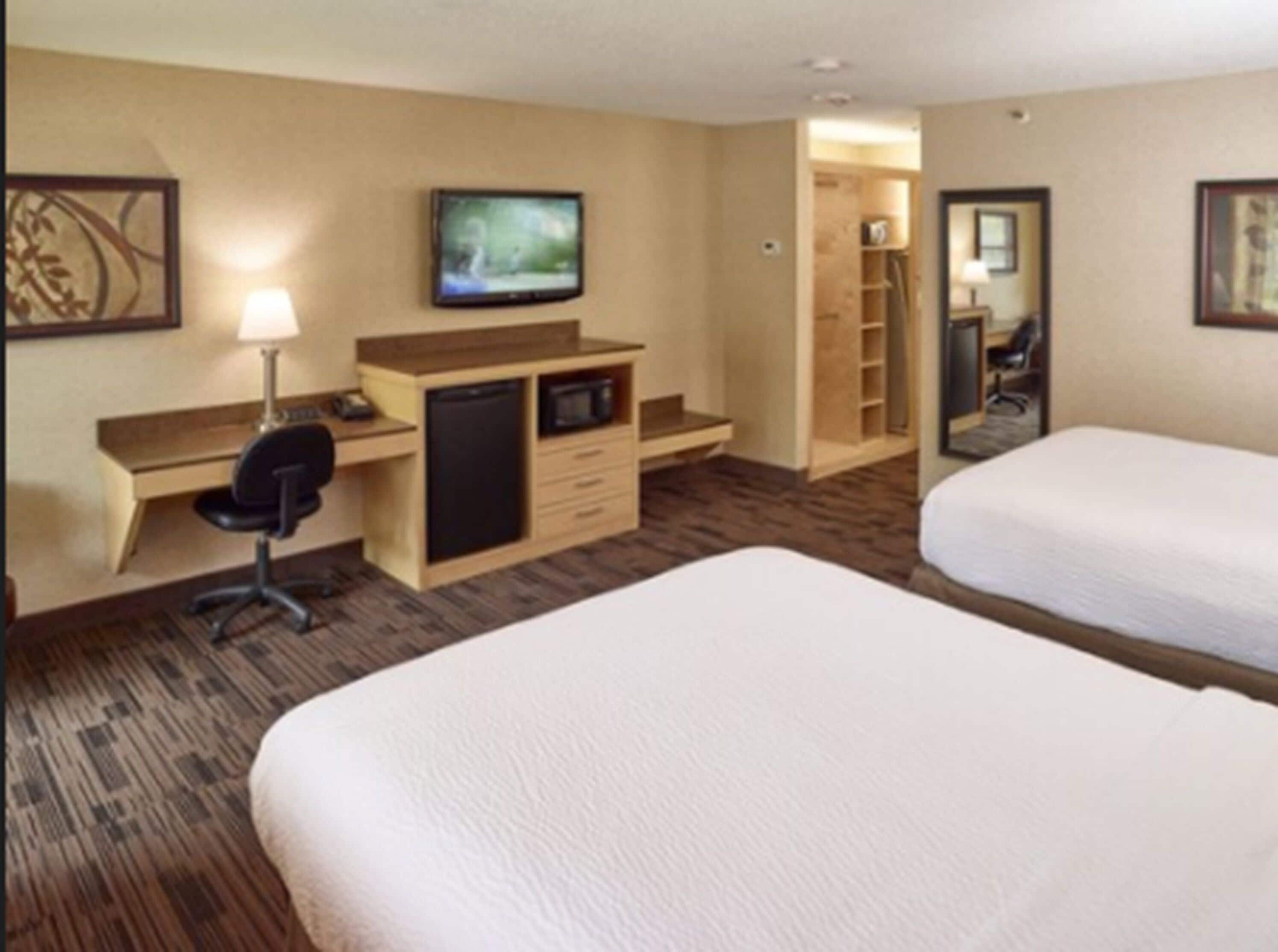 A hotel room with two beds, a desk with a chair, a television, a microwave, and a small kitchenette.
