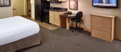 A hotel room with a bed, kitchenette, desk, chair, TV, and a dresser.