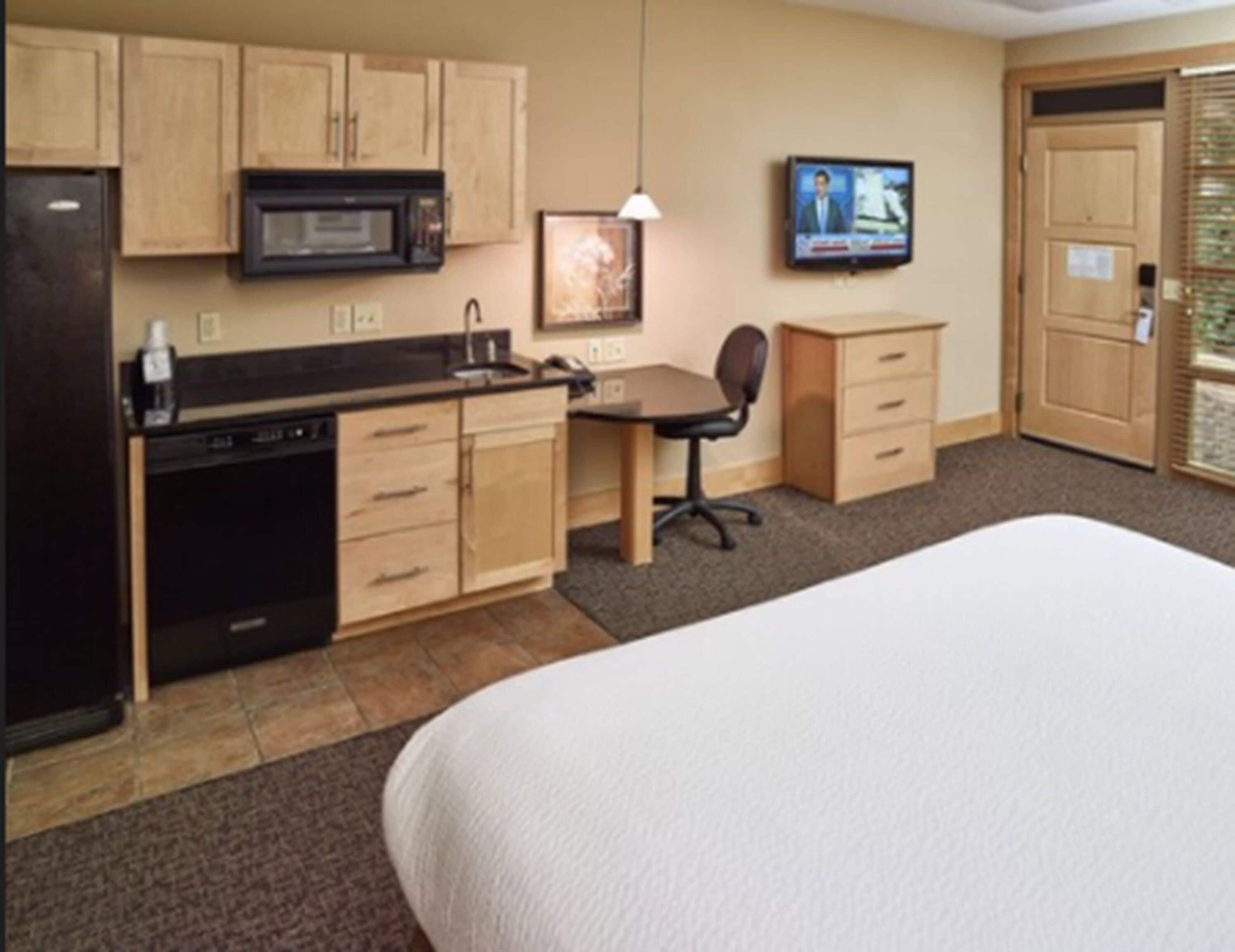 A hotel room with a bed, kitchenette, desk, chair, and a TV mounted on the wall.