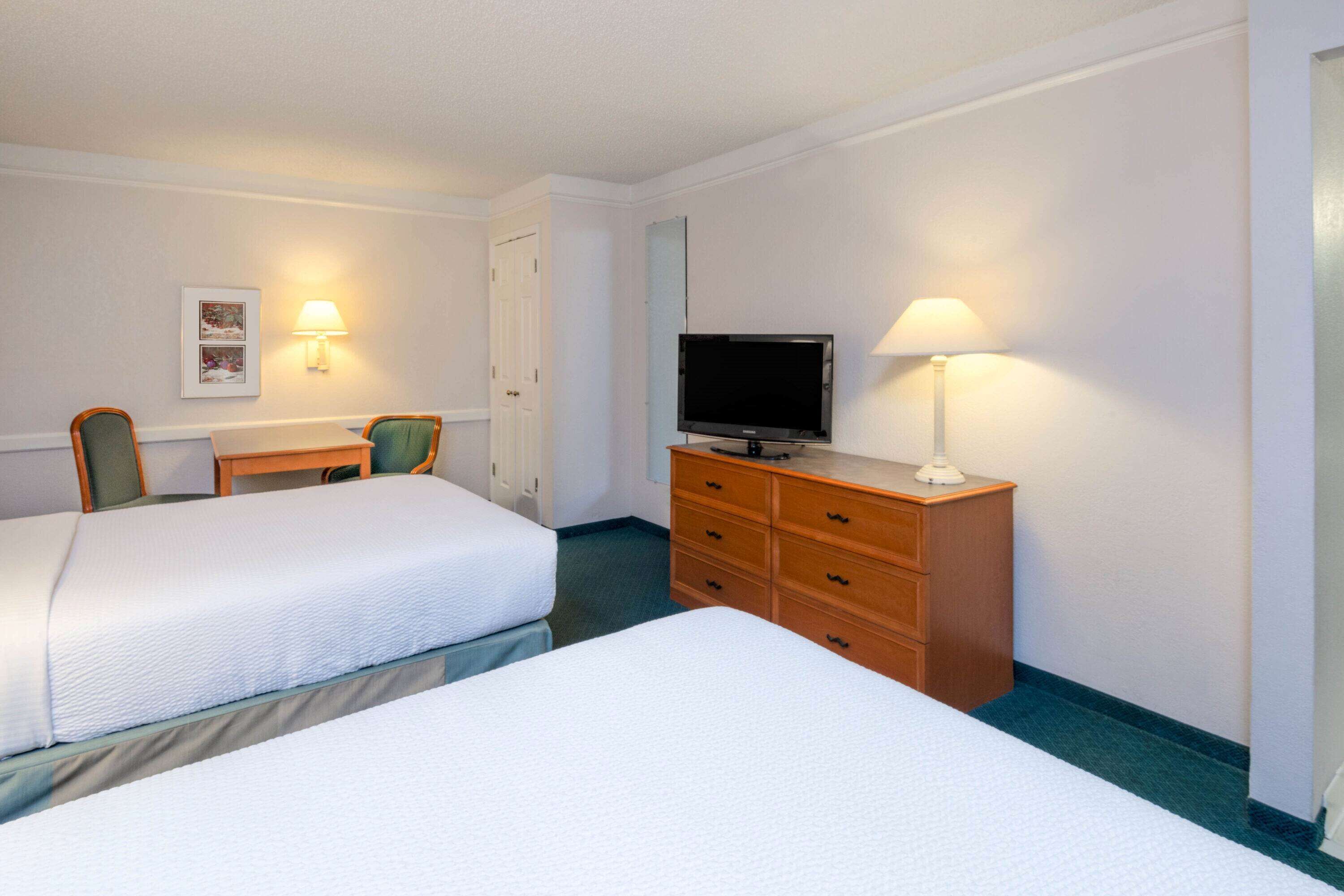 A hotel room with two beds, a television on a dresser, and two wall-mounted lamps.