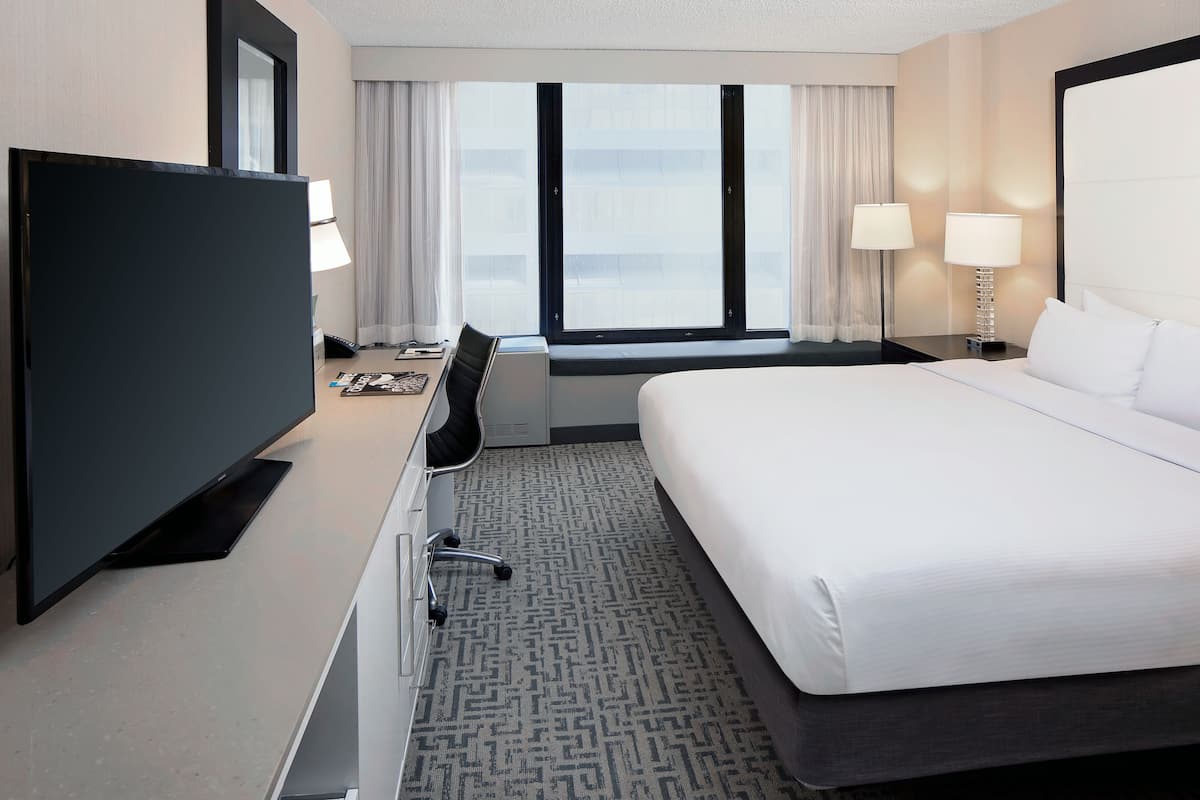 A hotel room with a large bed, a desk with a chair, a flat-screen TV, and a window with curtains.