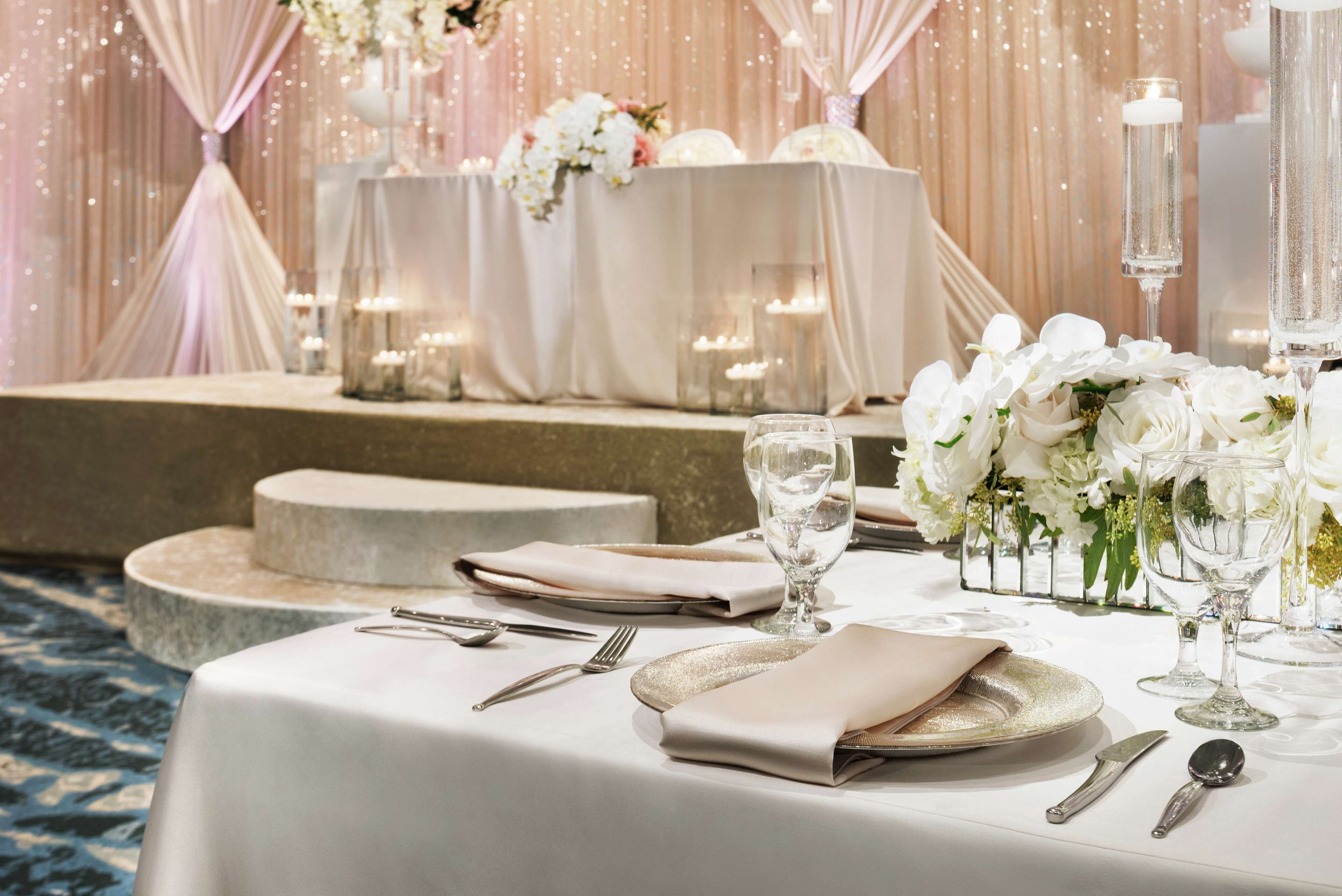 A banquet table set with white tablecloths, silverware, and glassware, with a backdrop of draped curtains and floral arrangements.