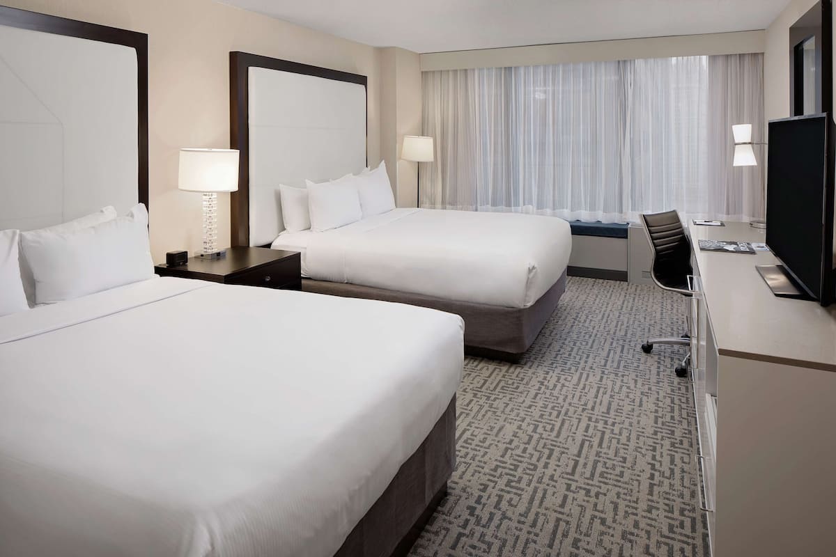 A hotel room with two beds, a desk with a chair, a television, and a large window with curtains.