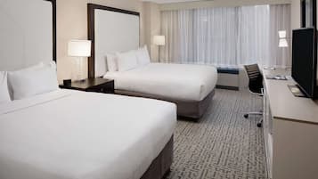 Habitación de hotel con dos camas, un escritorio con silla, televisor y un ventanal con cortinas.