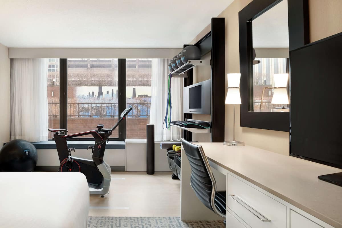 A hotel room with a bed, exercise equipment, a desk with a computer, a TV, and a large window with a city view.