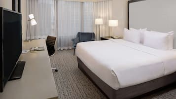 A hotel room with a large bed, a desk with a flat-screen TV, two chairs, a desk lamp, and a large window with sheer curtains.