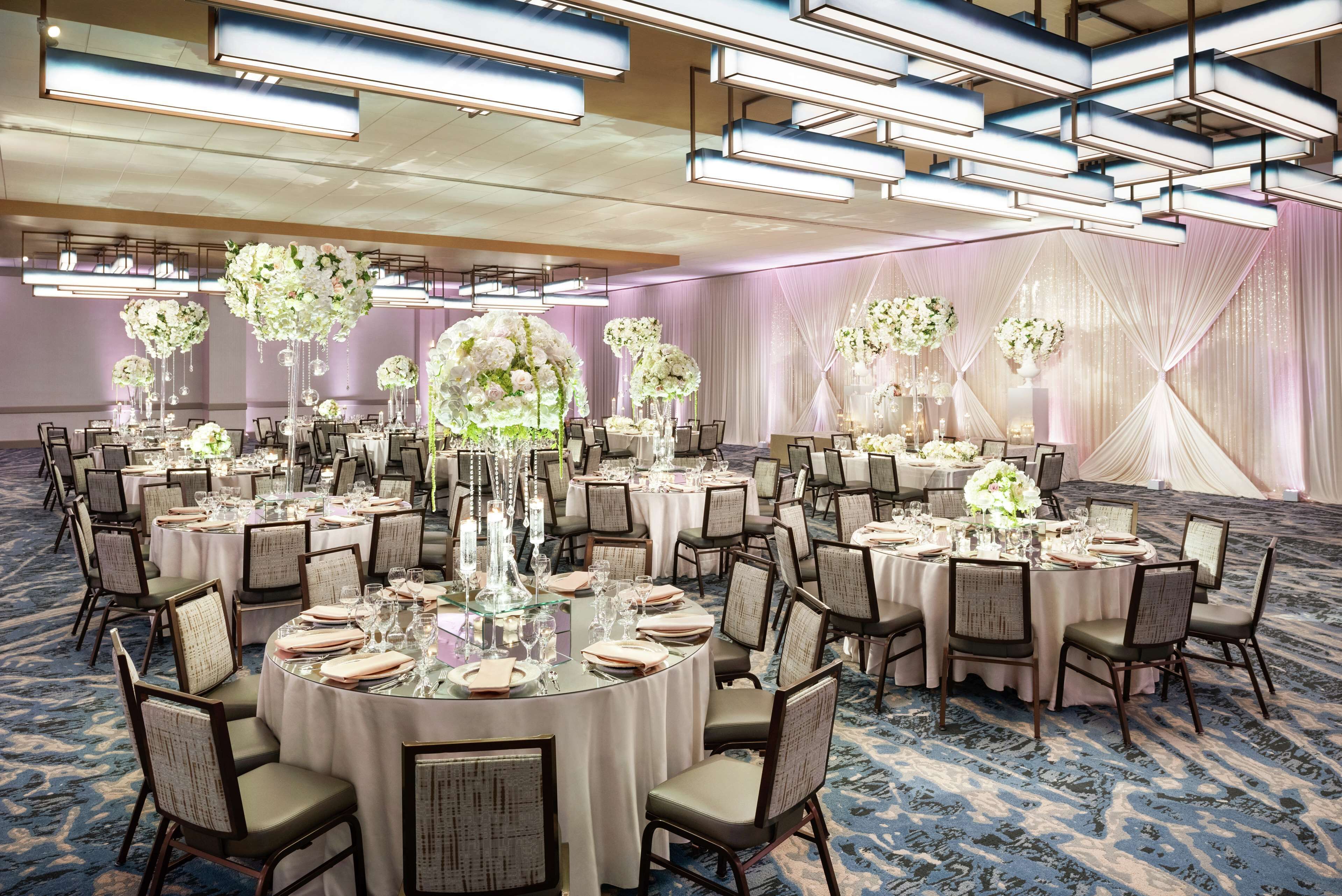 A banquet hall with round tables set for dining, featuring elegant floral centerpieces and a carpeted floor.