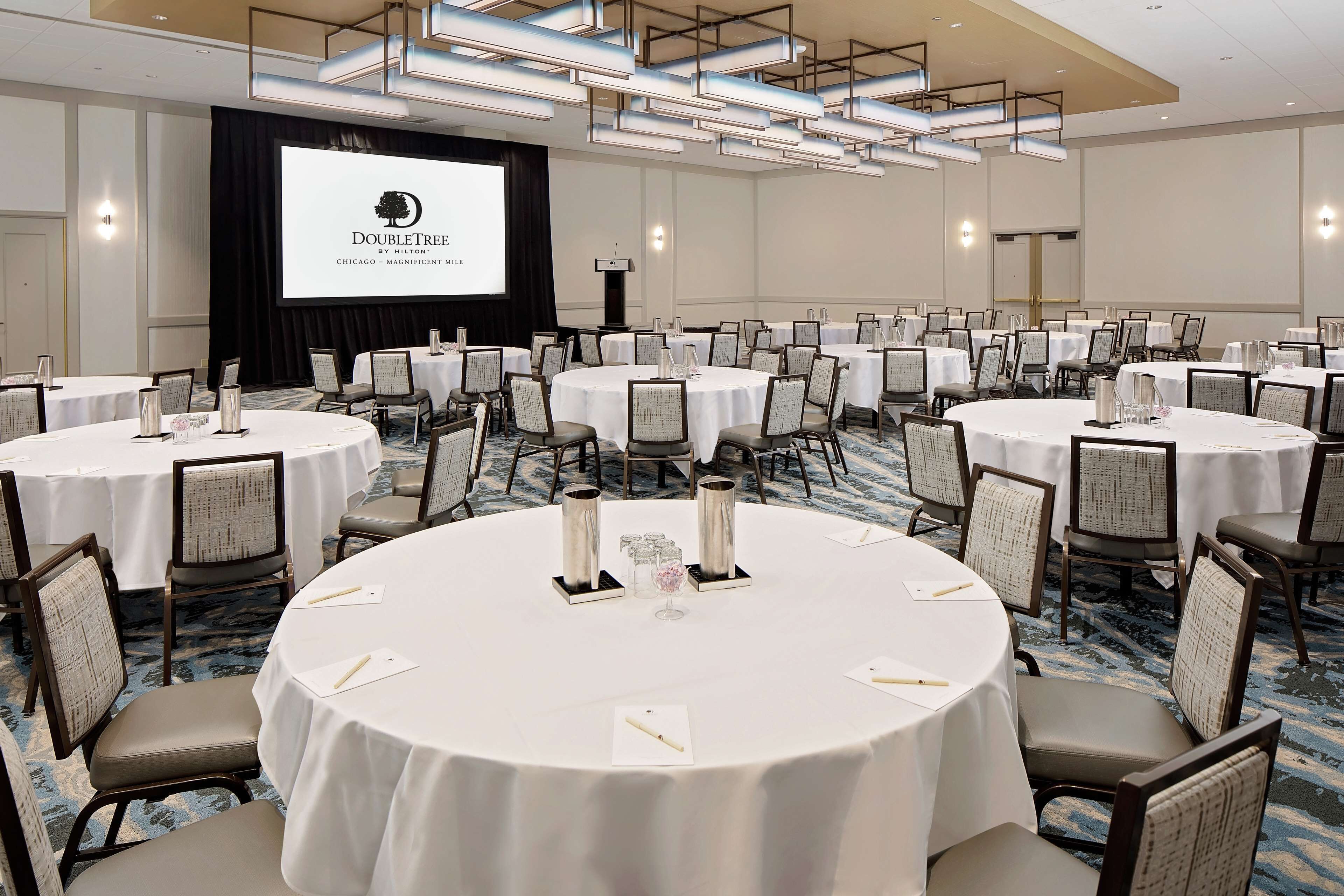 A conference room with multiple round tables, each with a white tablecloth and chairs, a projector screen displaying a presentation, and a carpeted floor.