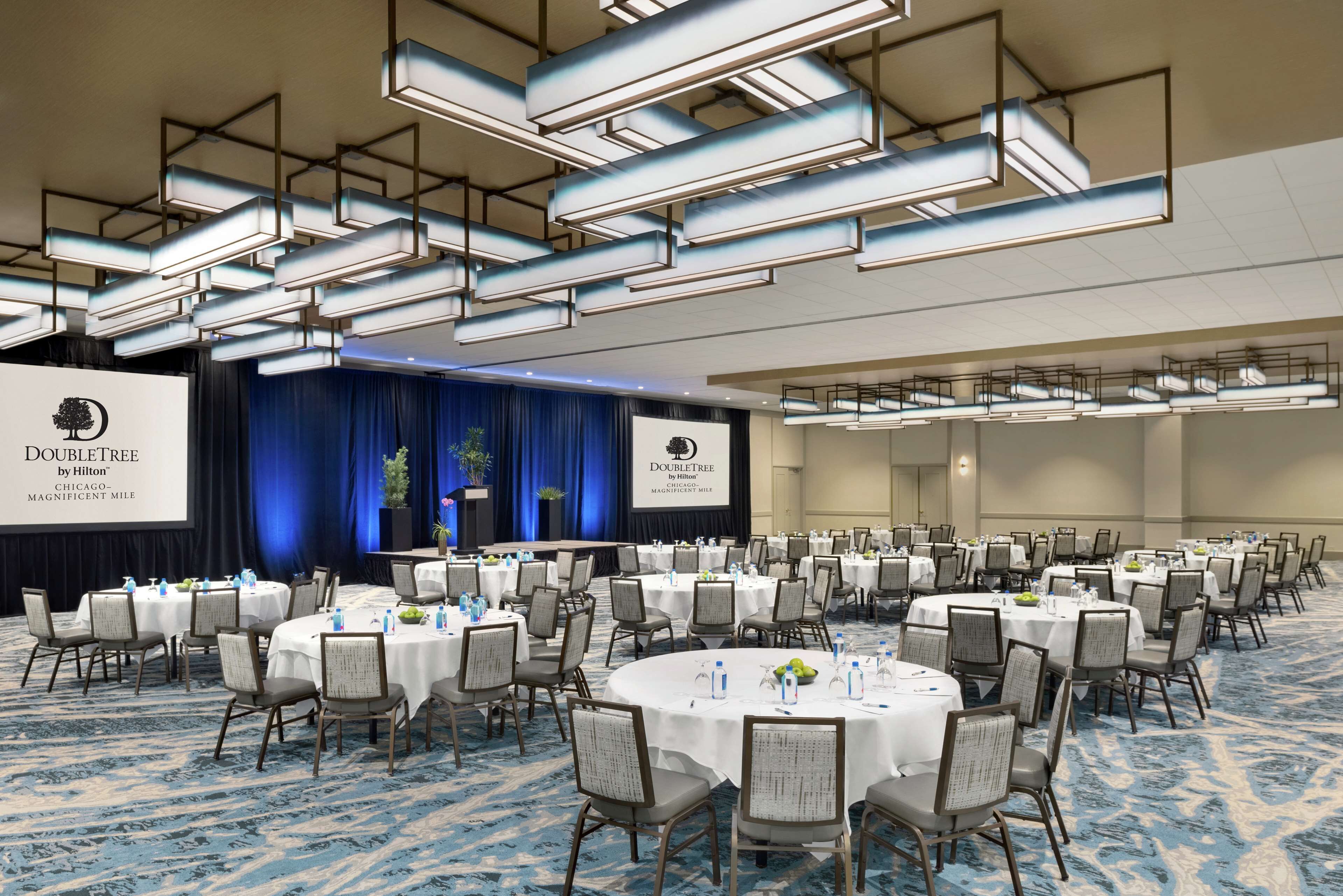 A conference room with round tables, chairs, and a stage with a presentation screen.