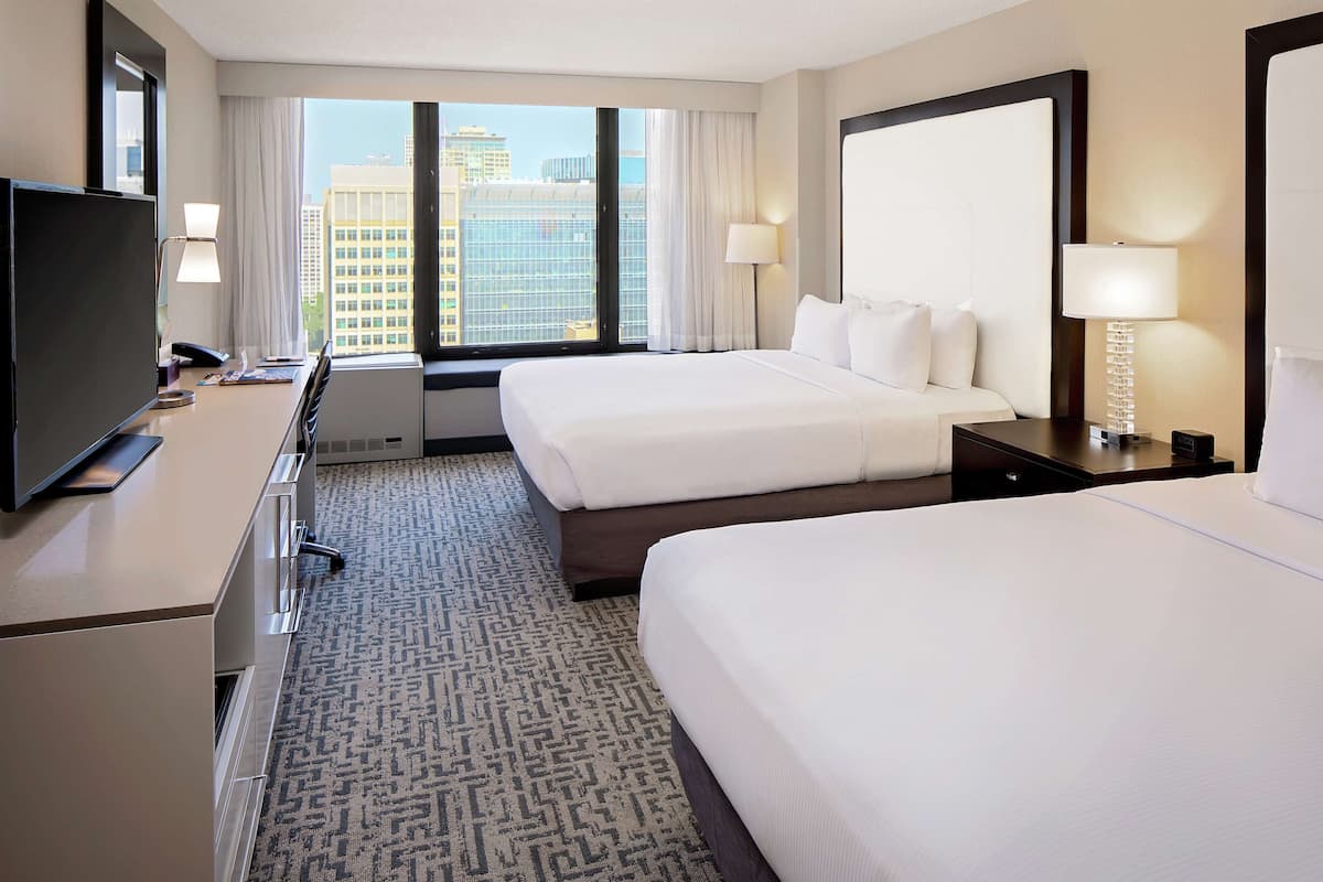 A hotel room with two beds, a TV, a desk, and a large window with a city view.