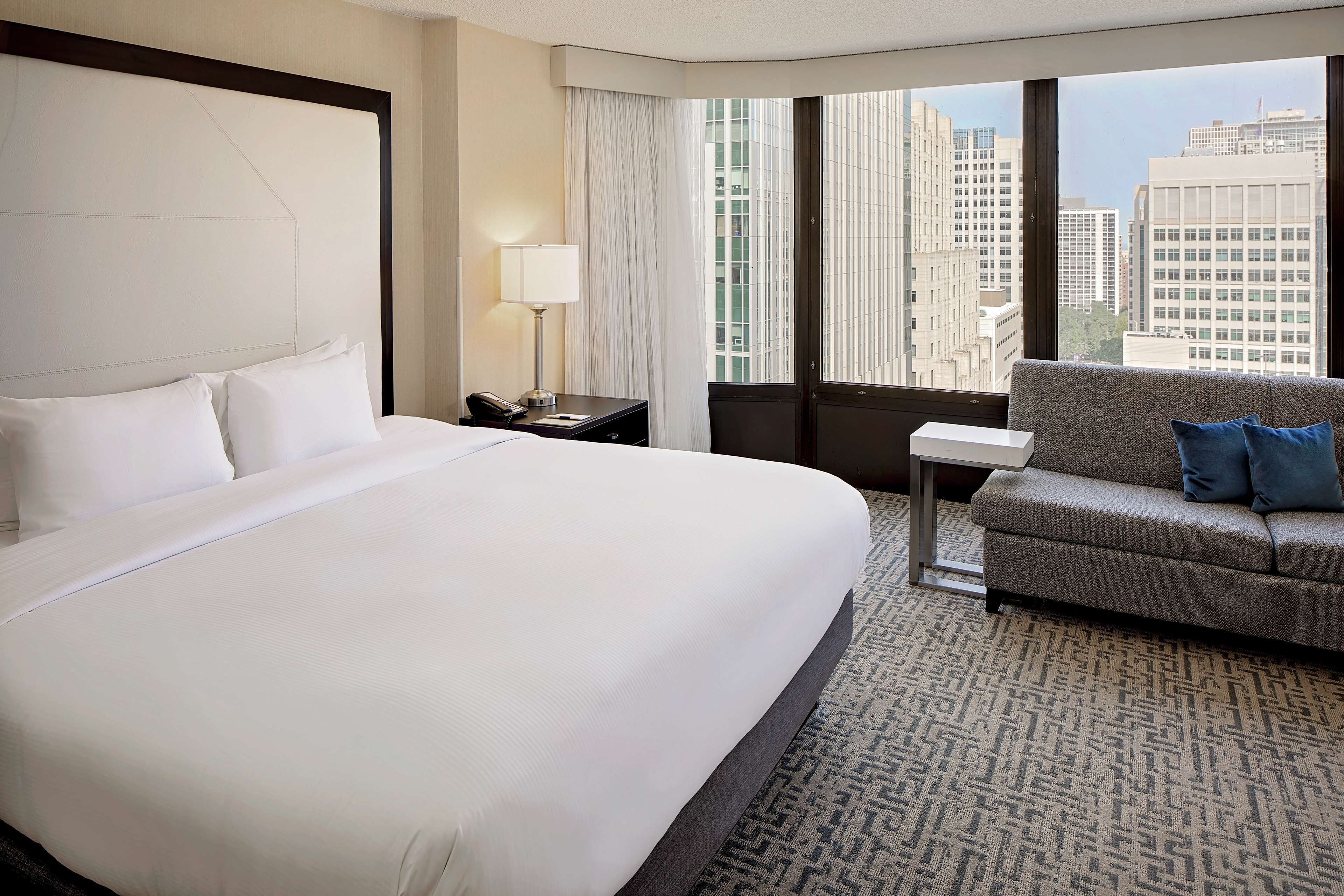 A hotel room with a large bed, a sofa, a nightstand with a lamp, and a view of tall buildings.