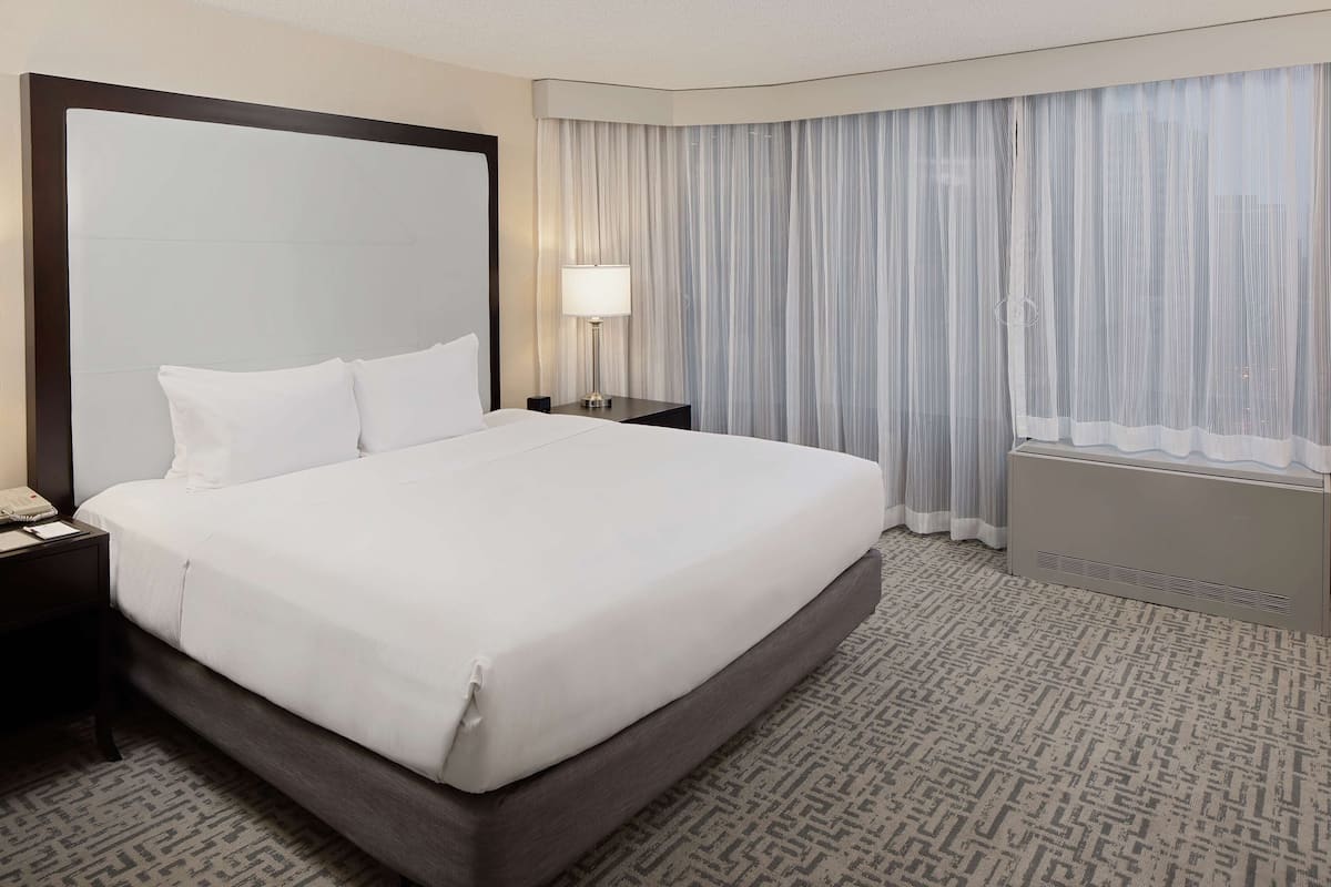 A hotel room with a large bed, a nightstand with a lamp, a window with curtains, and a radiator.
