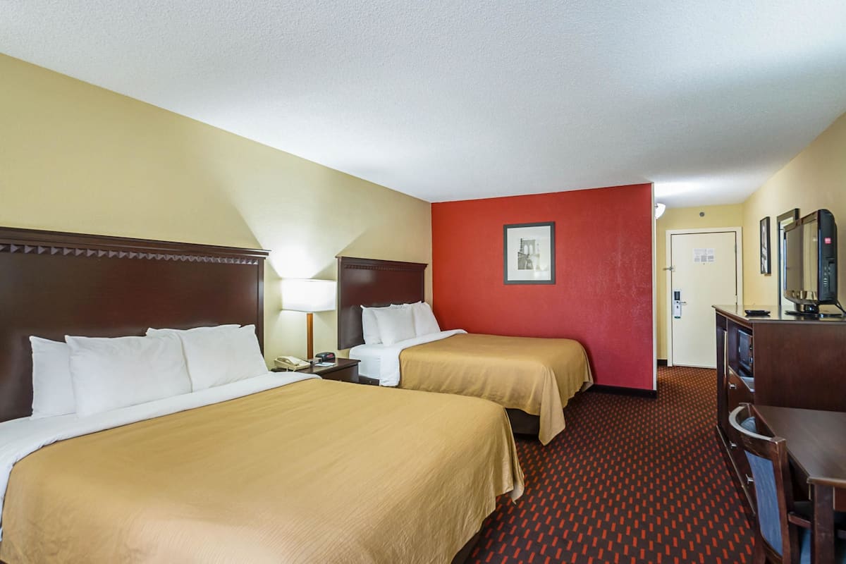 A hotel room with two beds, a red accent wall, a desk with a computer, and a door leading to a bathroom.
