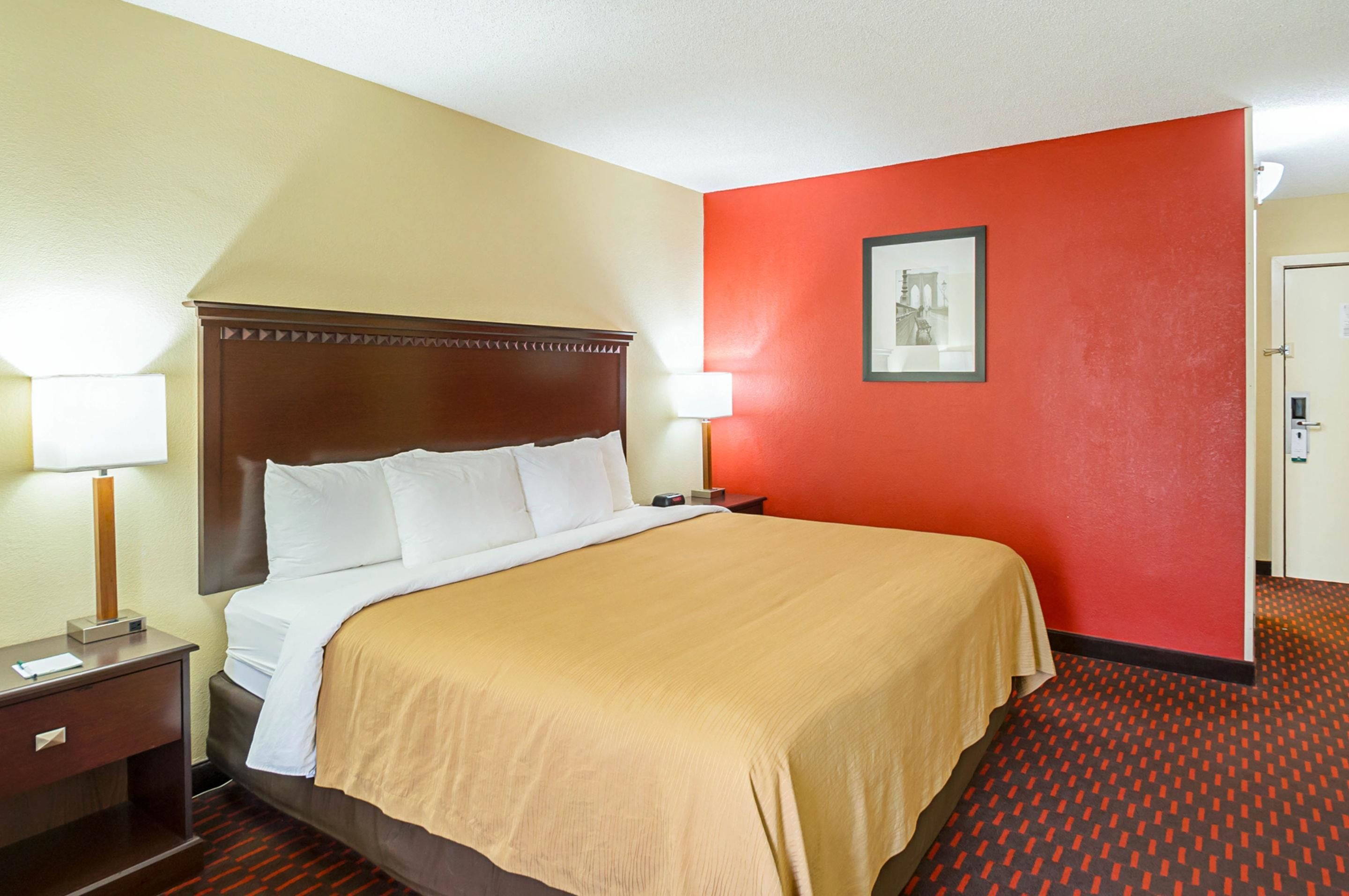 Habitación de hotel con una cama grande, una mesita de noche con lámpara, pared de acento roja y un cuadro enmarcado en la pared.