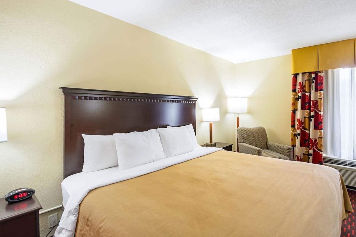 A hotel room with a large bed, a nightstand with a clock, a chair, and a lamp.