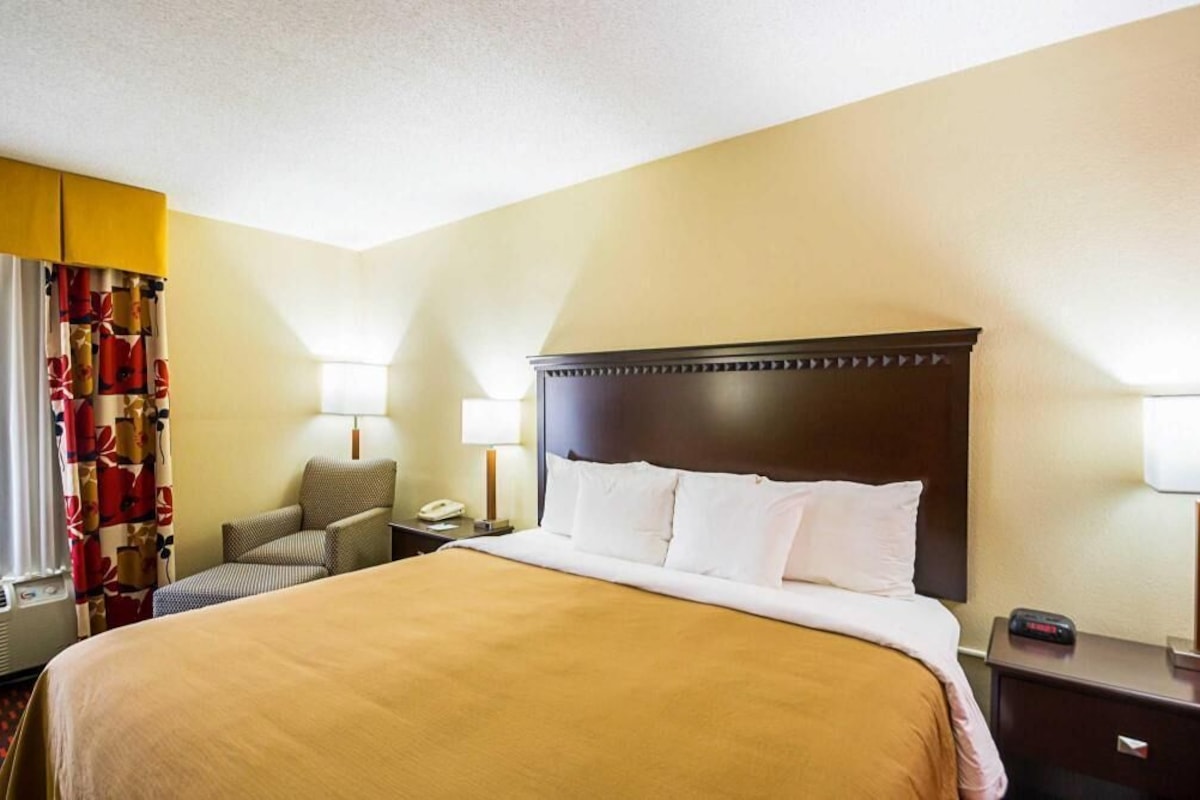 A hotel room with a large bed, two bedside lamps, a chair, a desk with a phone, and a window with curtains.
