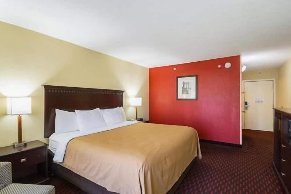 A hotel room with a bed, bedside tables, a red accent wall, and a framed picture.