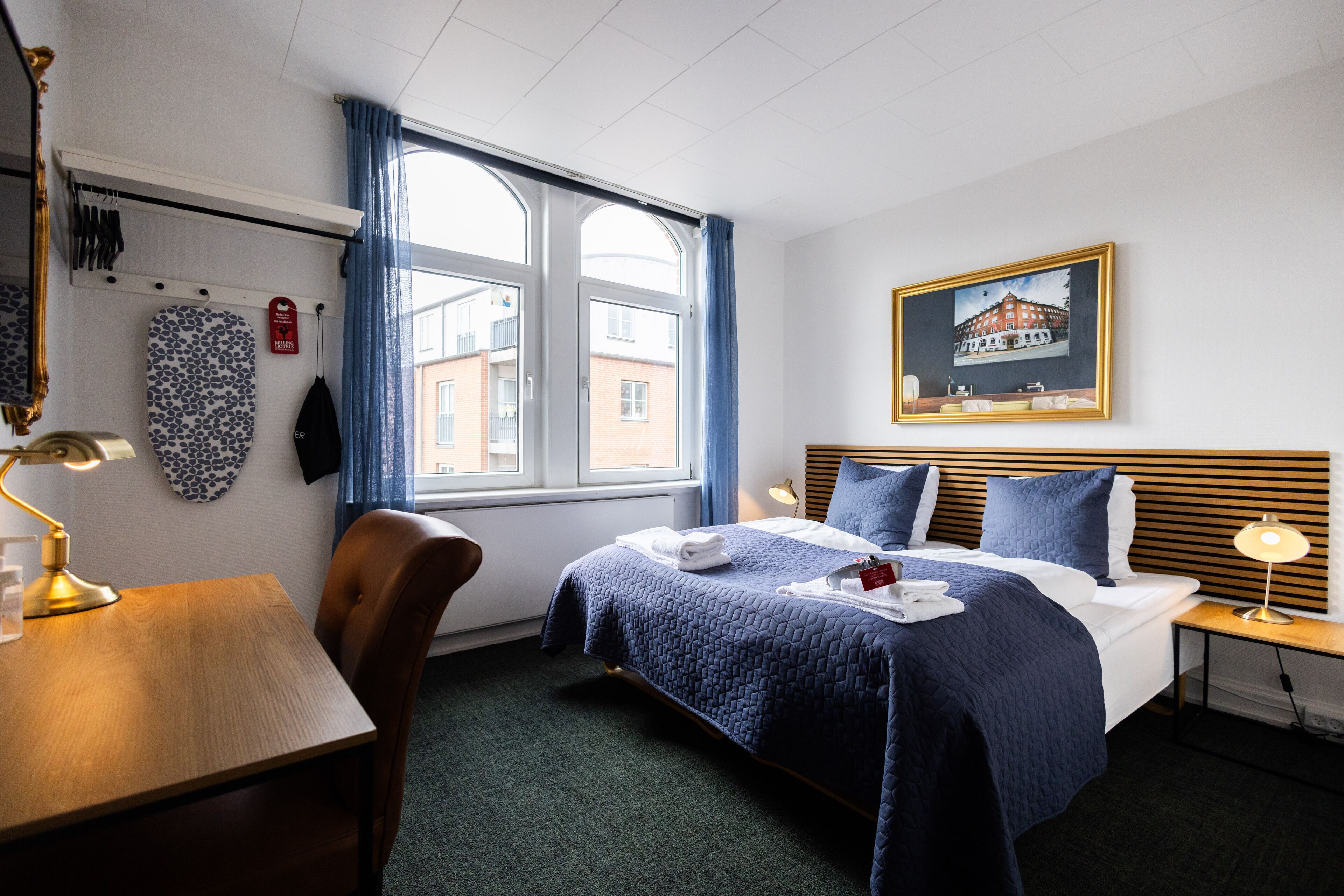 Hotellihuone, jossa on sänky, työpöytä, tuoli, ikkuna verhoilla, taulu seinällä ja lamppu.