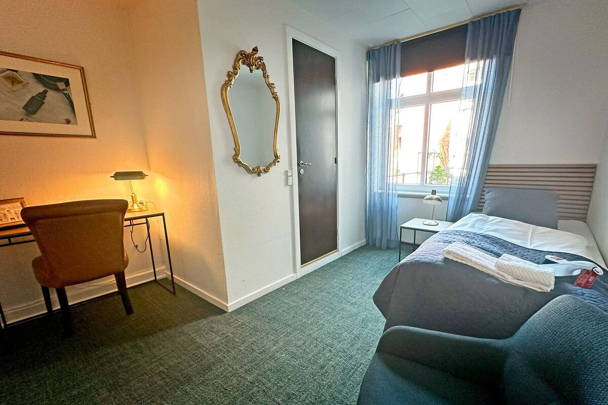 A hotel room with a bed, a desk with a lamp, a chair, a mirror, and a window with curtains.