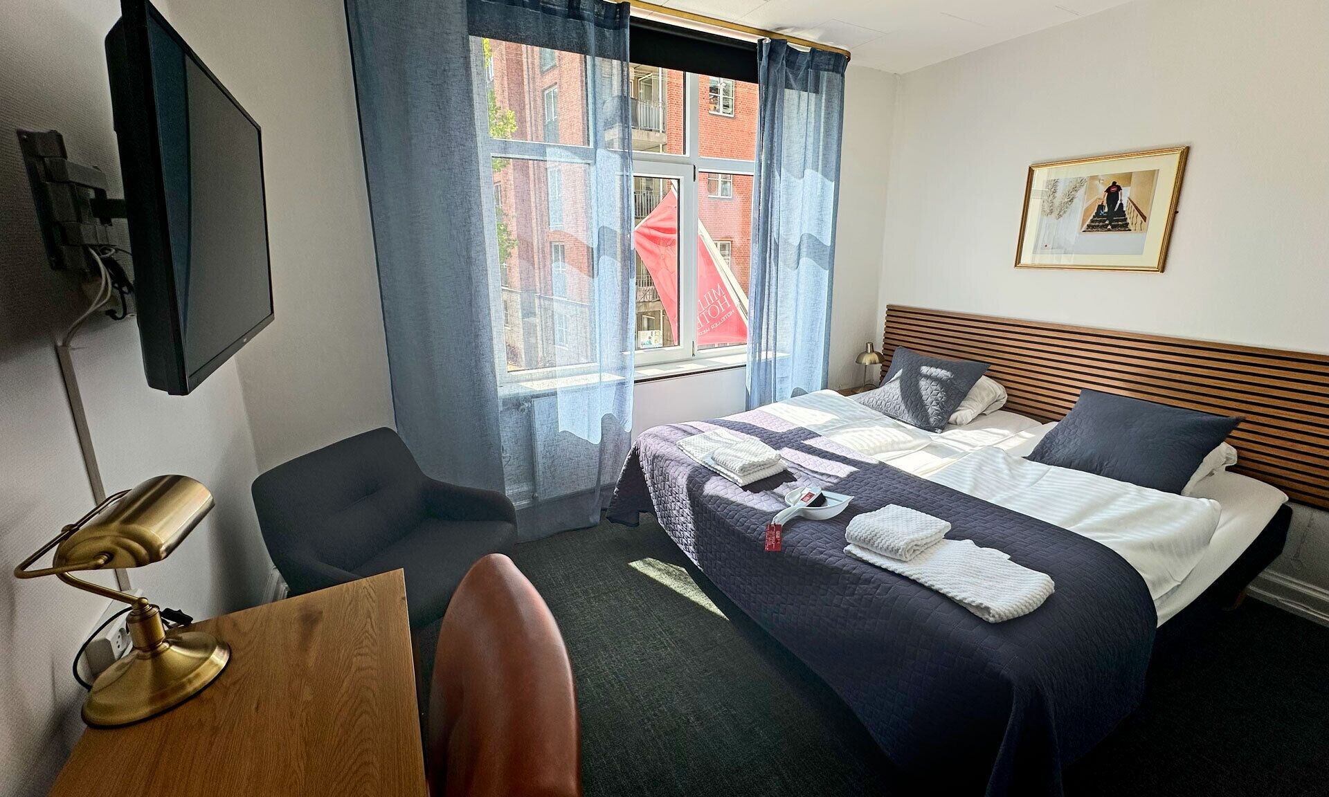 Une chambre d’hôtel avec un lit, un bureau, une chaise, une télévision et une fenêtre avec des rideaux.