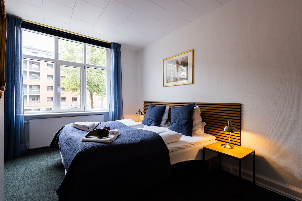 A hotel room with a large window, a bed with a blue bedspread, a bedside table with a lamp, and a framed picture on the wall.