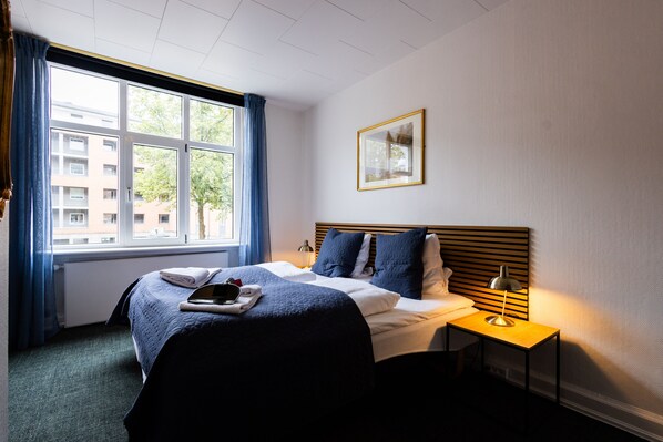 A hotel room with a large window, a bed with a blue bedspread, a bedside table with a lamp, and a framed picture on the wall.